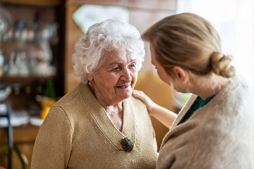 An elderly woman with white hair smiles warmly at a younger caregiver who is gently touching her shoulder in a supportive, comforting way.